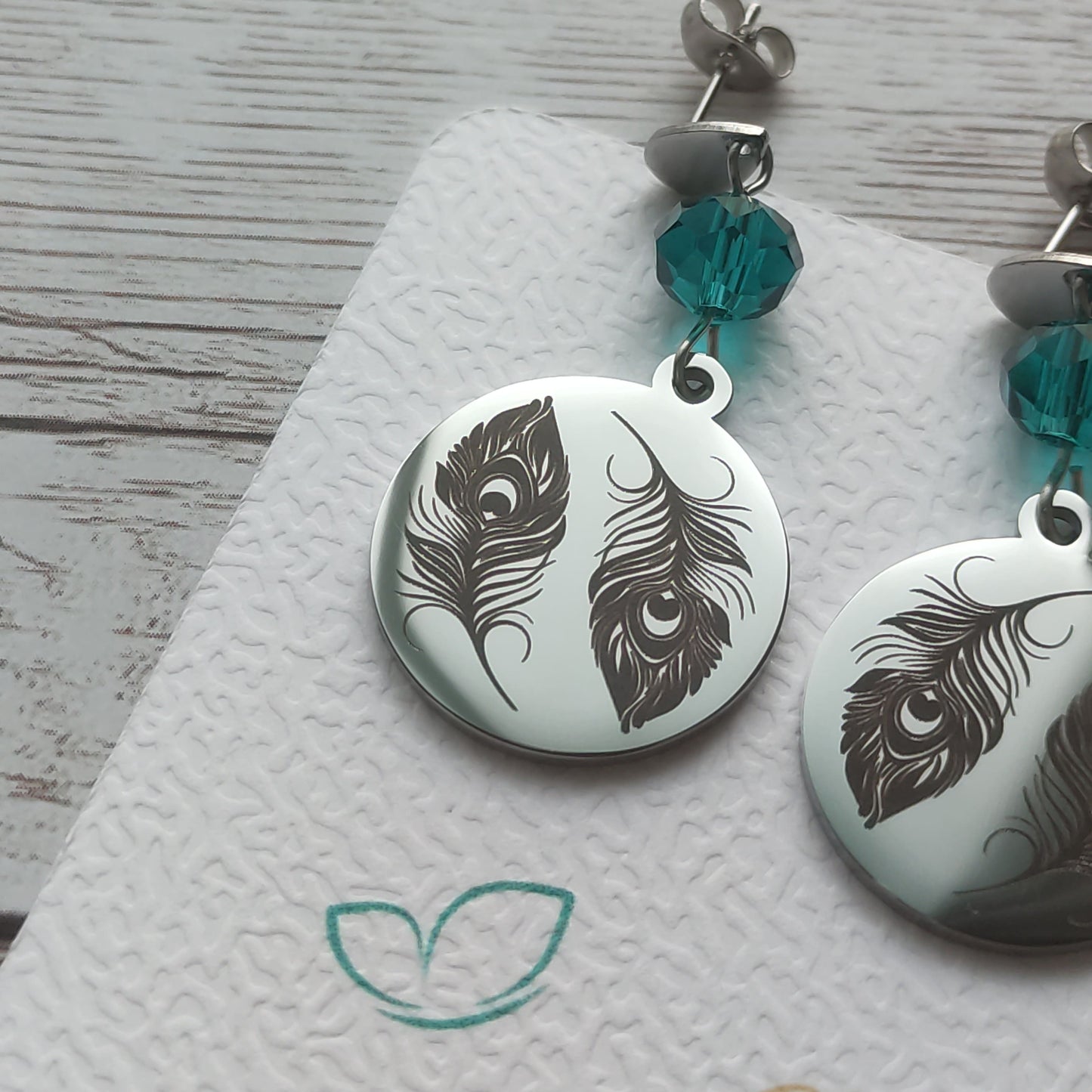 Boucles d'oreilles pendantes en acier inoxydable poli miroir avec gravure laser de deux plumes de paon et cristal à facettes.