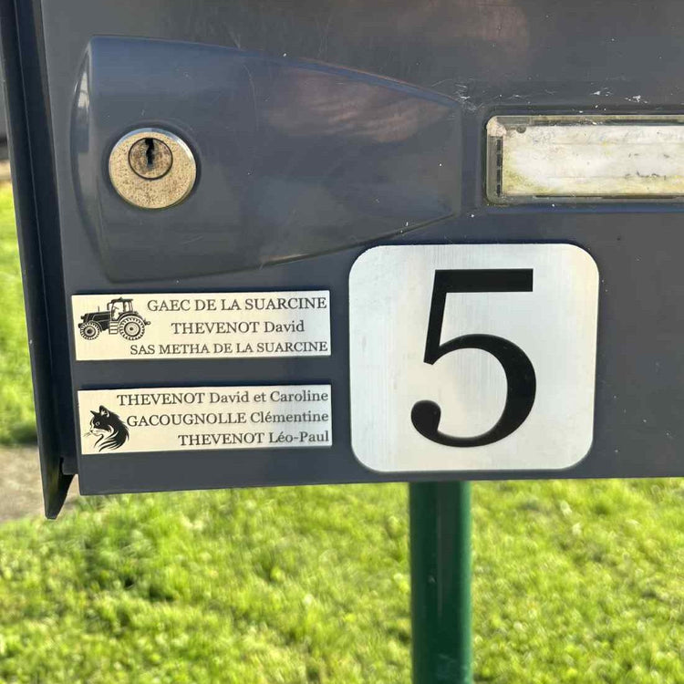 Gros plan sur une plaque acrylique argent brossé personnalisée avec gravure laser d'un tracteur et texte professionnel détaillé : "GAEC DE LA SUARCINE / THEVENOT David / SAS METHA DE LA SUARCINE".
