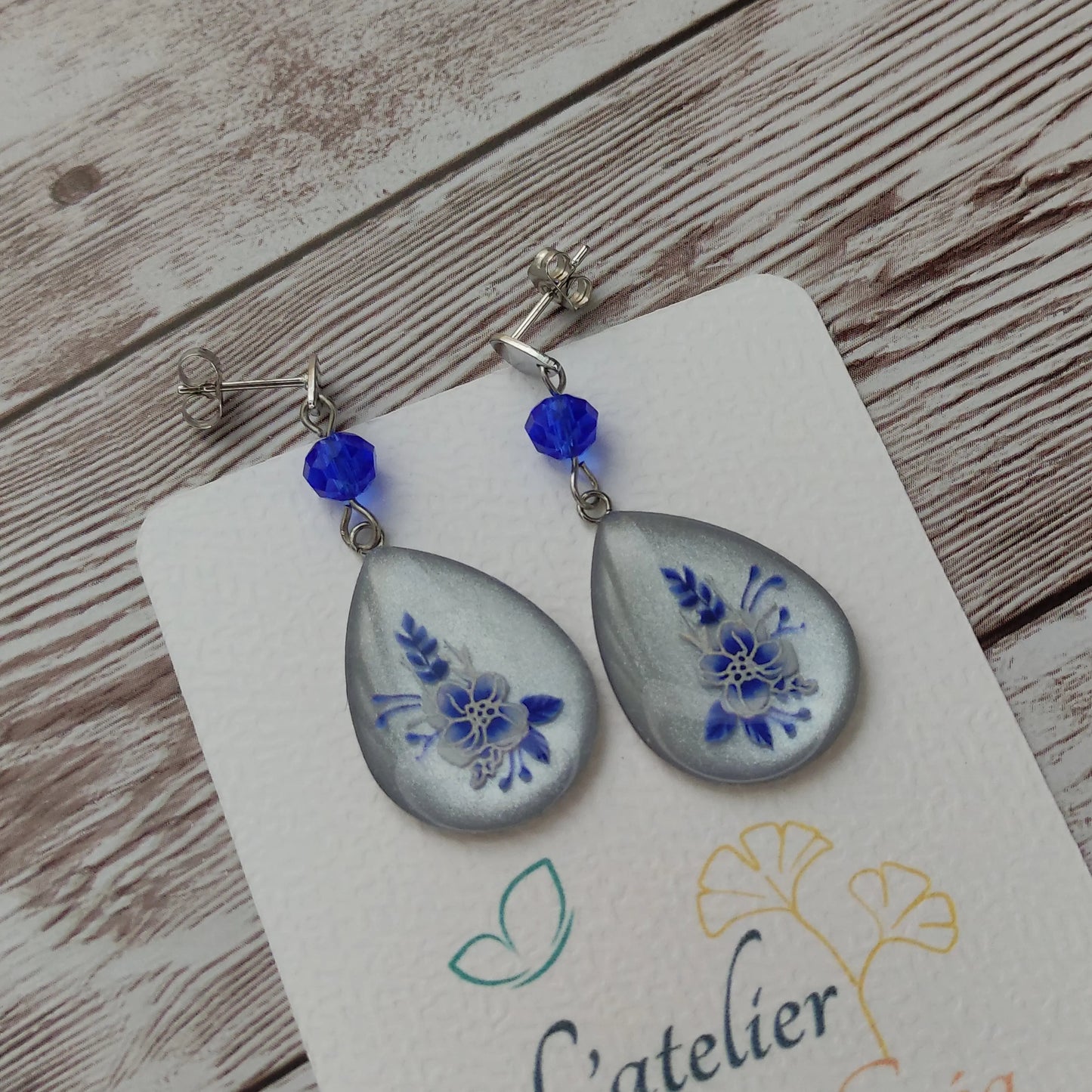 Boucles d'oreilles goutte fleurs résine bleues et blanches sur fond blanc nacré et clous en acier inoxydable.