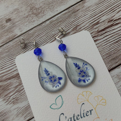 Boucles d'oreilles goutte fleurs résine bleues et blanches sur fond blanc nacré et clous en acier inoxydable.