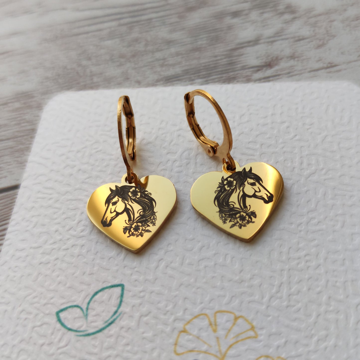 Paire de boucles d'oreilles dormeuses en acier inoxydable avec pendentif coeur poli miroir gravé d'une tête de cheval et de fleurs.
