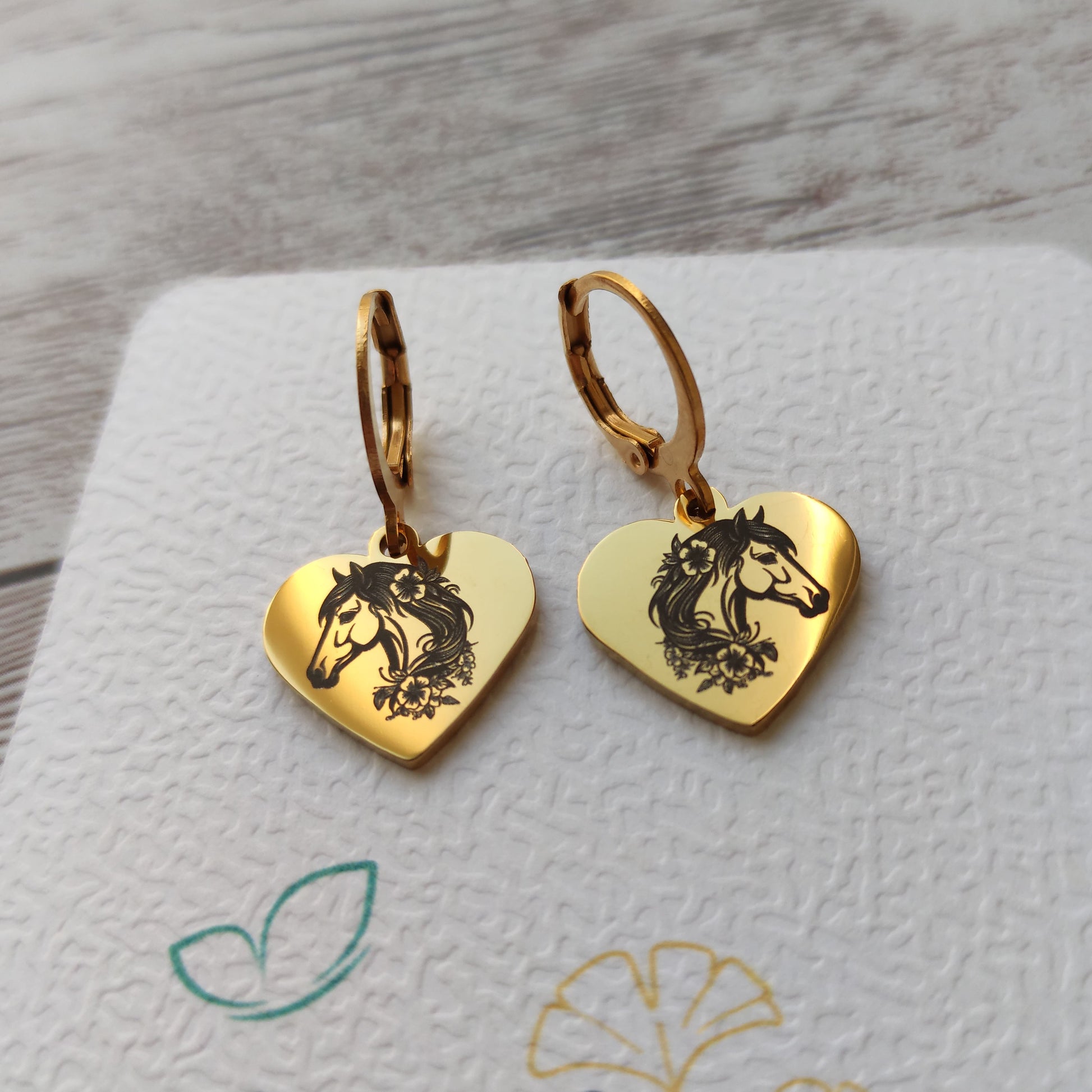 Paire de boucles d'oreilles dormeuses en acier inoxydable avec pendentif coeur poli miroir gravé d'une tête de cheval et de fleurs.
