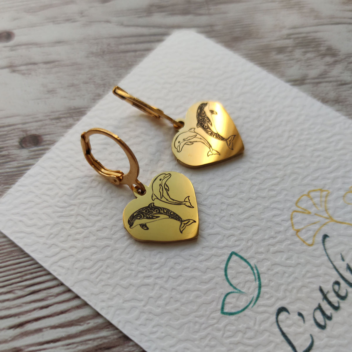Paire de boucles d'oreilles dormeuses en acier inoxydable avec pendentif coeur gravé de deux dauphins, finition poli miroir.