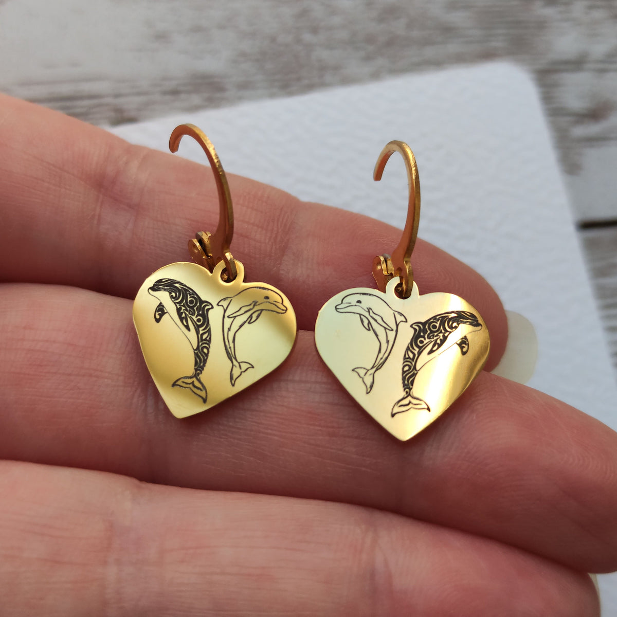 Paire de boucles d'oreilles dormeuses en acier inoxydable avec pendentif coeur gravé de deux dauphins, finition poli miroir.