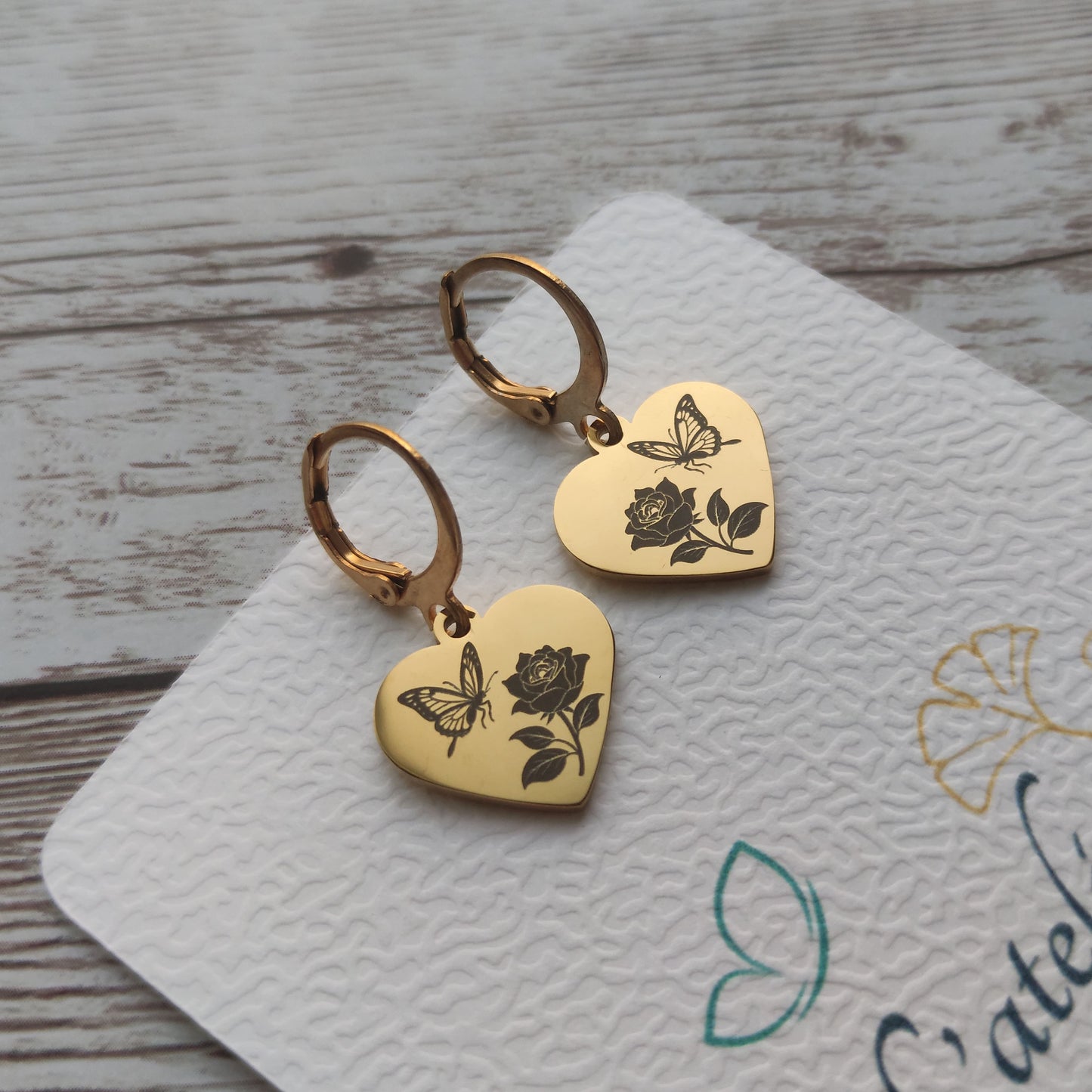 Paire de dormeuses en acier inoxydable avec pendentif coeur poli miroir gravé d'un papillon et d'une rose sur fond blanc.