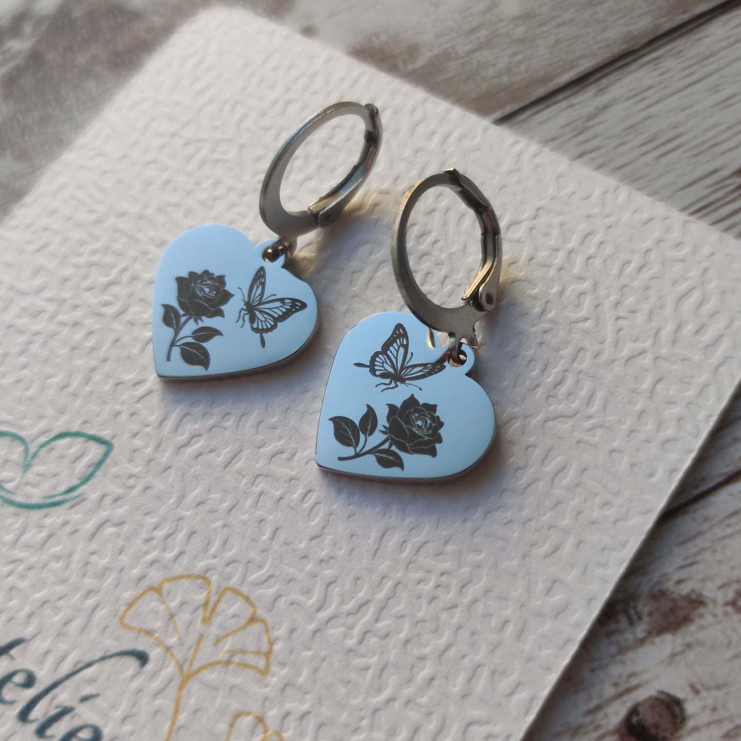 Paire de dormeuses en acier inoxydable avec pendentif coeur poli miroir gravé d'un papillon et d'une rose sur fond blanc.