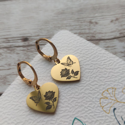 Paire de dormeuses en acier inoxydable avec pendentif coeur poli miroir gravé d'un papillon et d'une rose sur fond blanc.