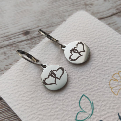 Boucles d'oreilles dormeuses en acier inoxydable poli miroir avec gravure laser de deux coeurs entrelacés sur médaillon rond.