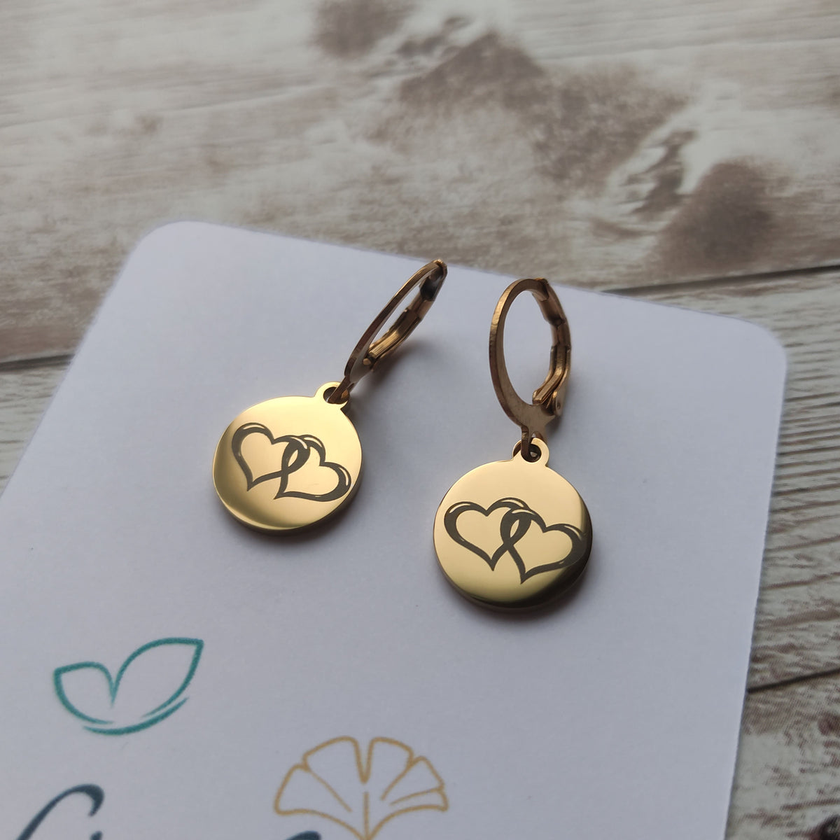 Boucles d'oreilles dormeuses en acier inoxydable poli miroir avec gravure laser de deux coeurs entrelacés sur médaillon rond.