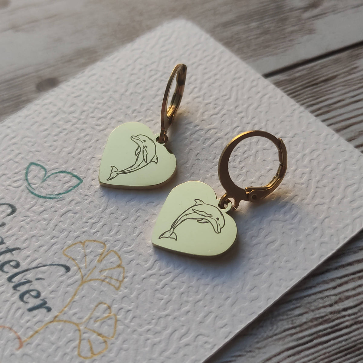 Paire de boucles d'oreilles dormeuses dauphin acier inoxydable avec pendentif coeur poli miroir et gravure laser de précision.