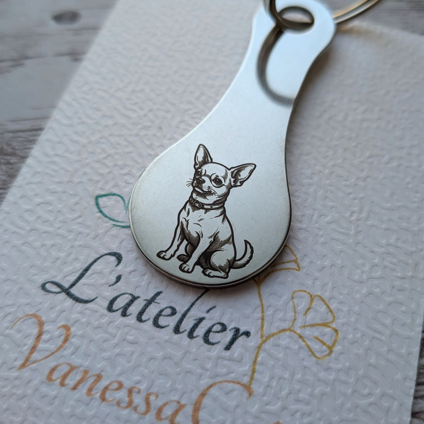 Porte-clés jeton de caddie en acier inoxydable gravé d'un petit chien de race Chihuahua assis, posé sur une carte de présentation de L'atelier Vanessa Créa.
