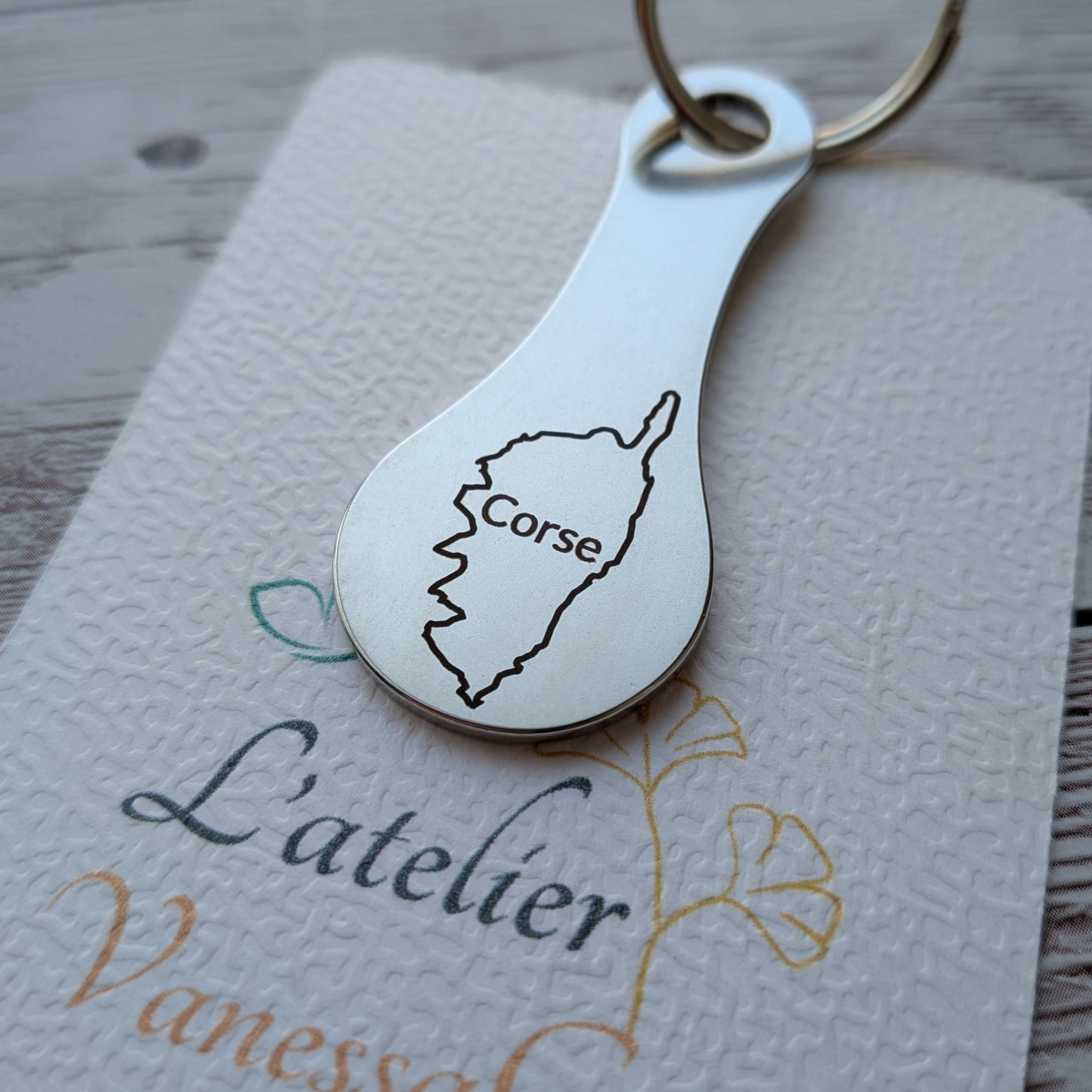 Porte-clés jeton de caddie en acier inoxydable gravé de la carte de l'île de la Corse avec son nom, posé sur le carton texturé de L'atelier Vanessa Créa.