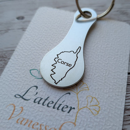 Porte-clés jeton de caddie en acier inoxydable gravé de la carte de l'île de la Corse avec son nom, posé sur le carton texturé de L'atelier Vanessa Créa.
