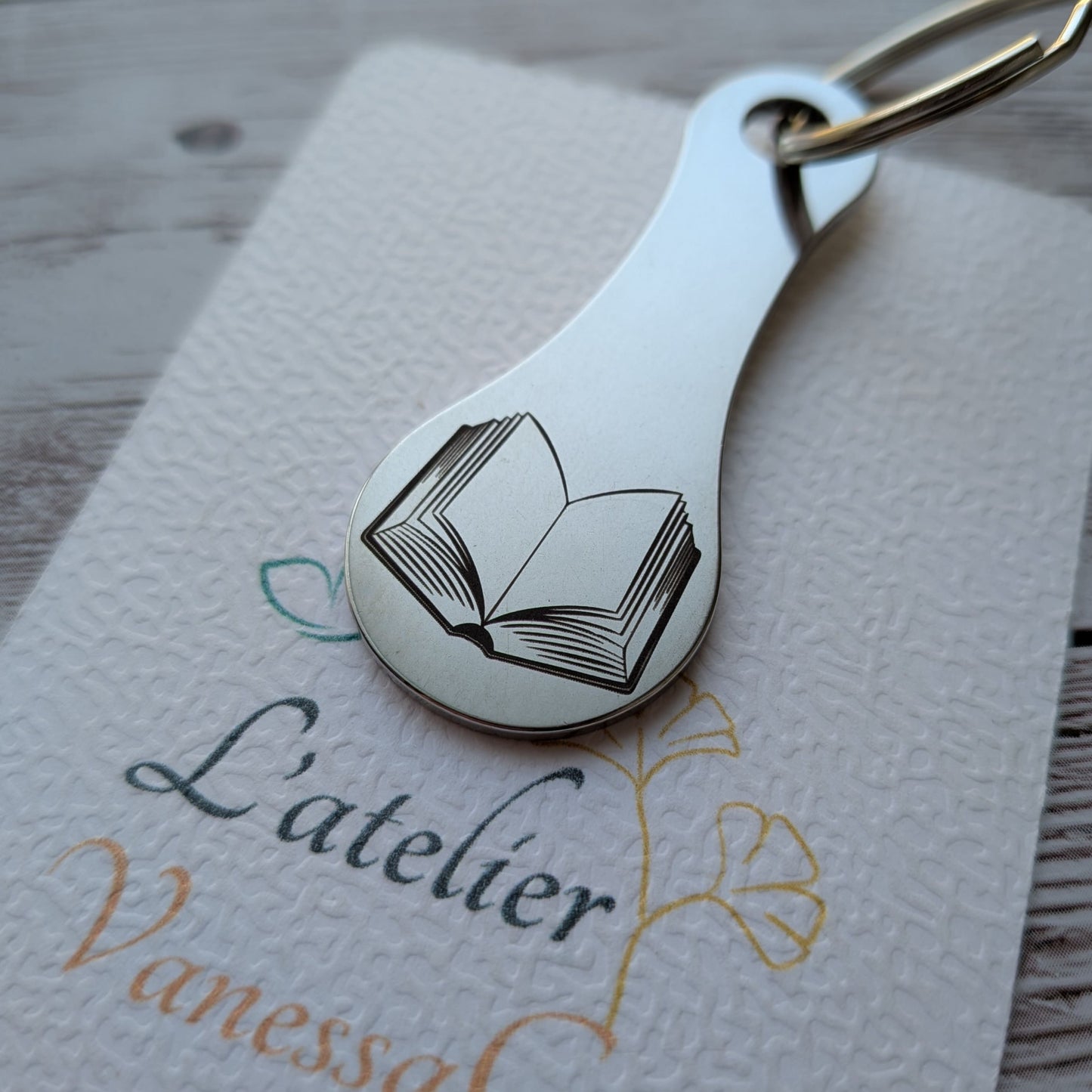 Porte-clés jeton de caddie en acier inoxydable avec une gravure détaillée d'un livre ouvert, présenté sur le papier texturé de L'atelier Vanessa Créa.