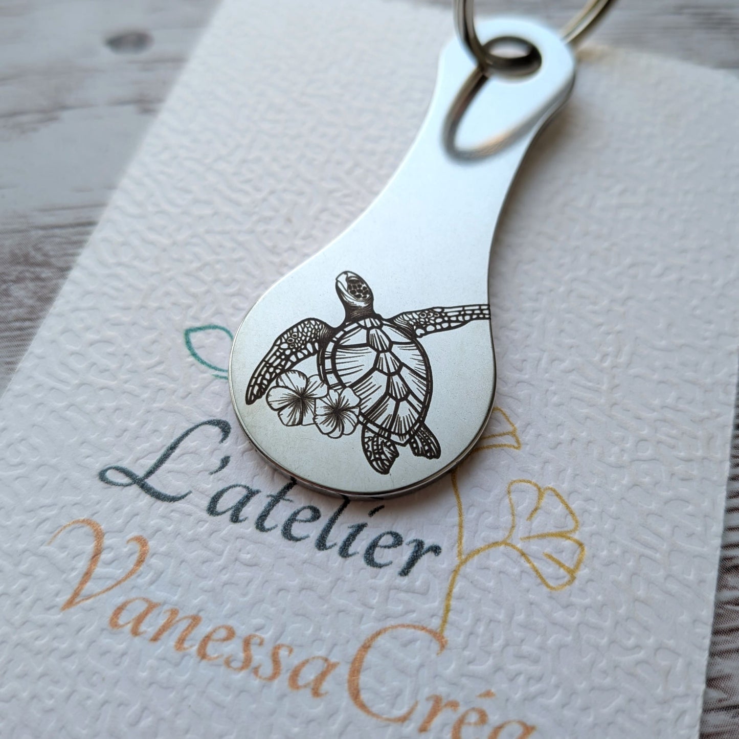 Porte-clés jeton de caddie en acier inoxydable gravé d'une tortue de mer stylisée accompagnée de fleurs tropicales, présenté dans L'atelier Vanessa Créa.