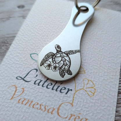 Porte-clés jeton de caddie en acier inoxydable gravé d'une tortue de mer stylisée accompagnée de fleurs tropicales, présenté dans L'atelier Vanessa Créa.