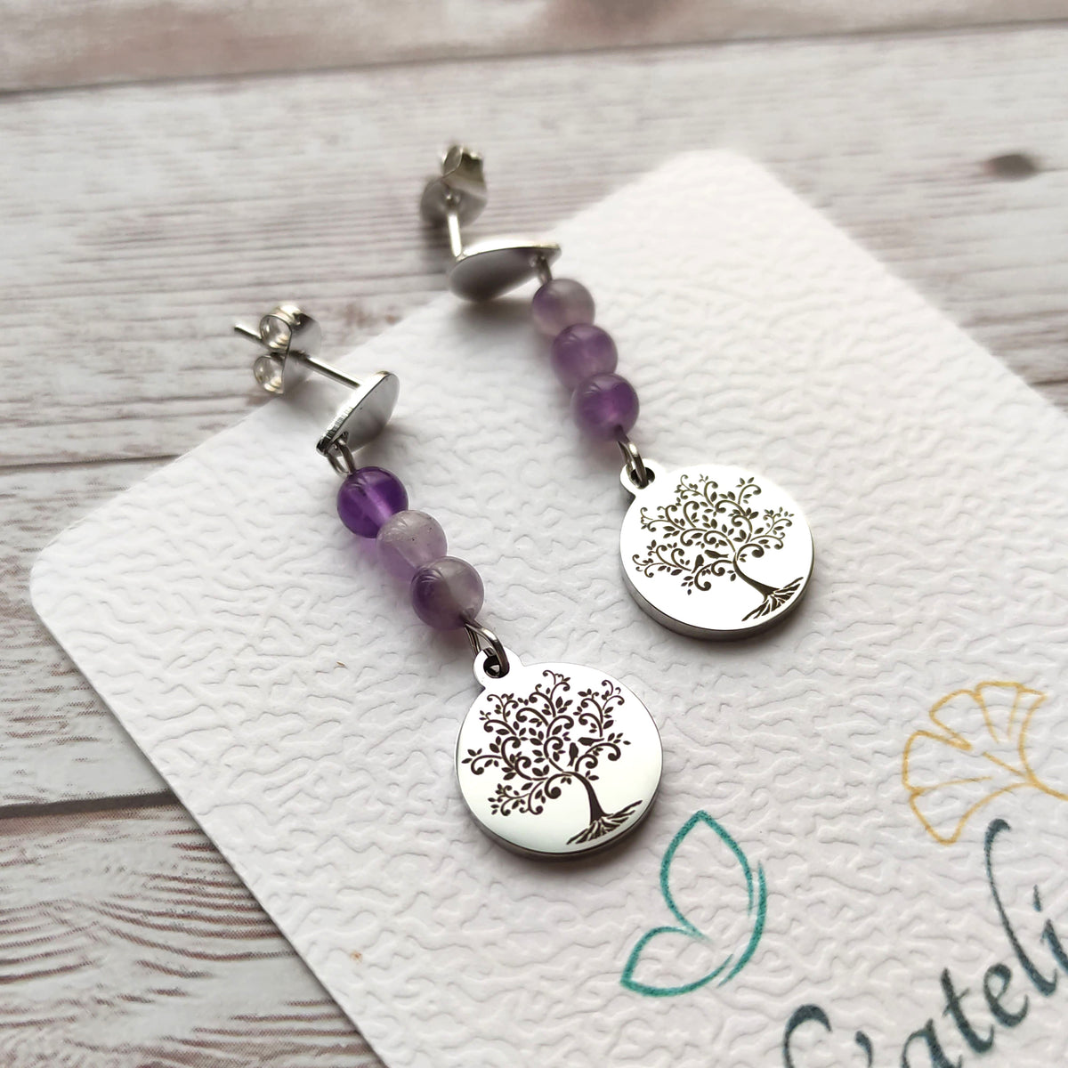Paire de boucles d'oreilles pendantes avec motif Arbre de Vie gravé et perles d'améthyste sur carte de présentation L'atelier Vanessa Créa.