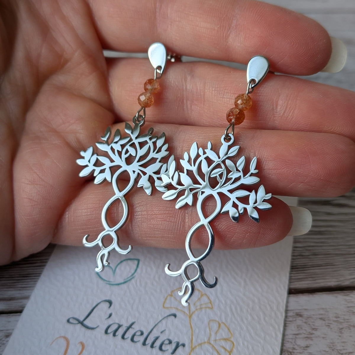 Paire de boucles d'oreilles arbre de vie argentées en acier inoxydable tenue en main.