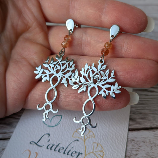 Paire de boucles d'oreilles arbre de vie argentées en acier inoxydable tenue en main.