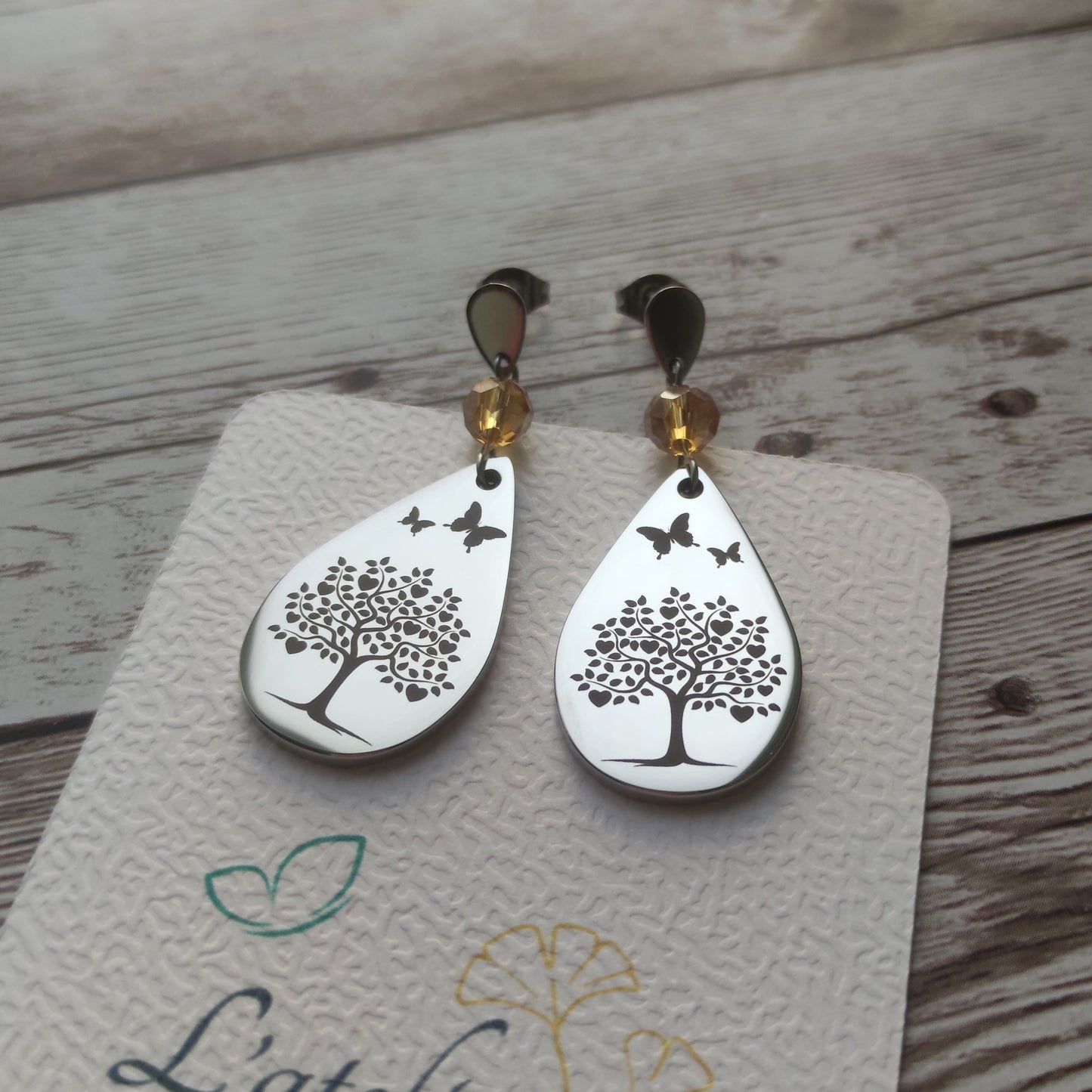Boucles d'oreilles pendantes en acier inoxydable poli miroir avec gravure laser d'un arbre de vie, cœurs et papillons, surmontées d'un cristal à facettes.