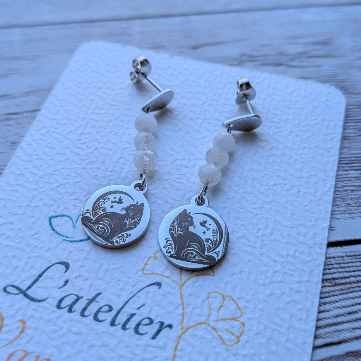 Paire de boucles d'oreilles pendantes avec motif chat gravé et perles naturelles sur carte de présentation L'atelier Vanessa Créa.