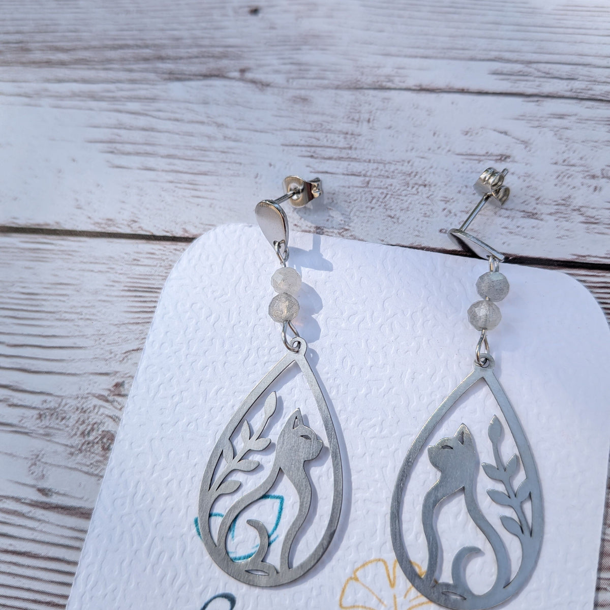 Paire de boucles d'oreilles artisanales motif chat et feuillage ajouré en acier inoxydable et labradorite sur support de présentation.