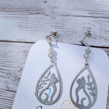 Paire de boucles d'oreilles artisanales motif chat et feuillage ajouré en acier inoxydable et labradorite sur support de présentation.