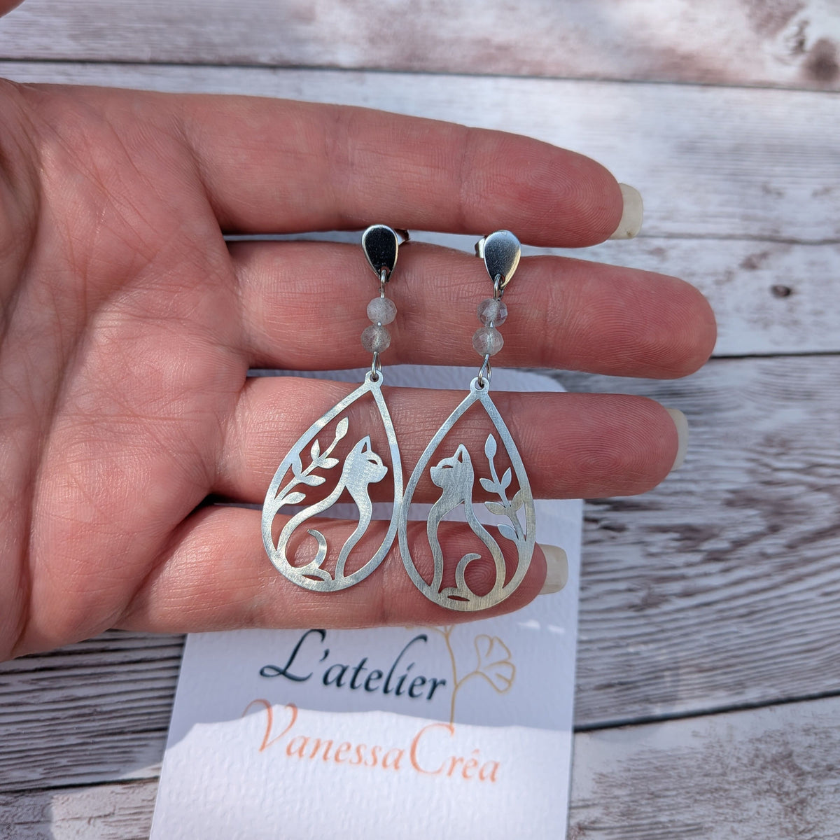 Boucles d'oreilles chat et feuillage ajouré en acier inoxydable avec perles de labradorite, présentées sur la main avec la carte de l'atelier Vanessa Créa.