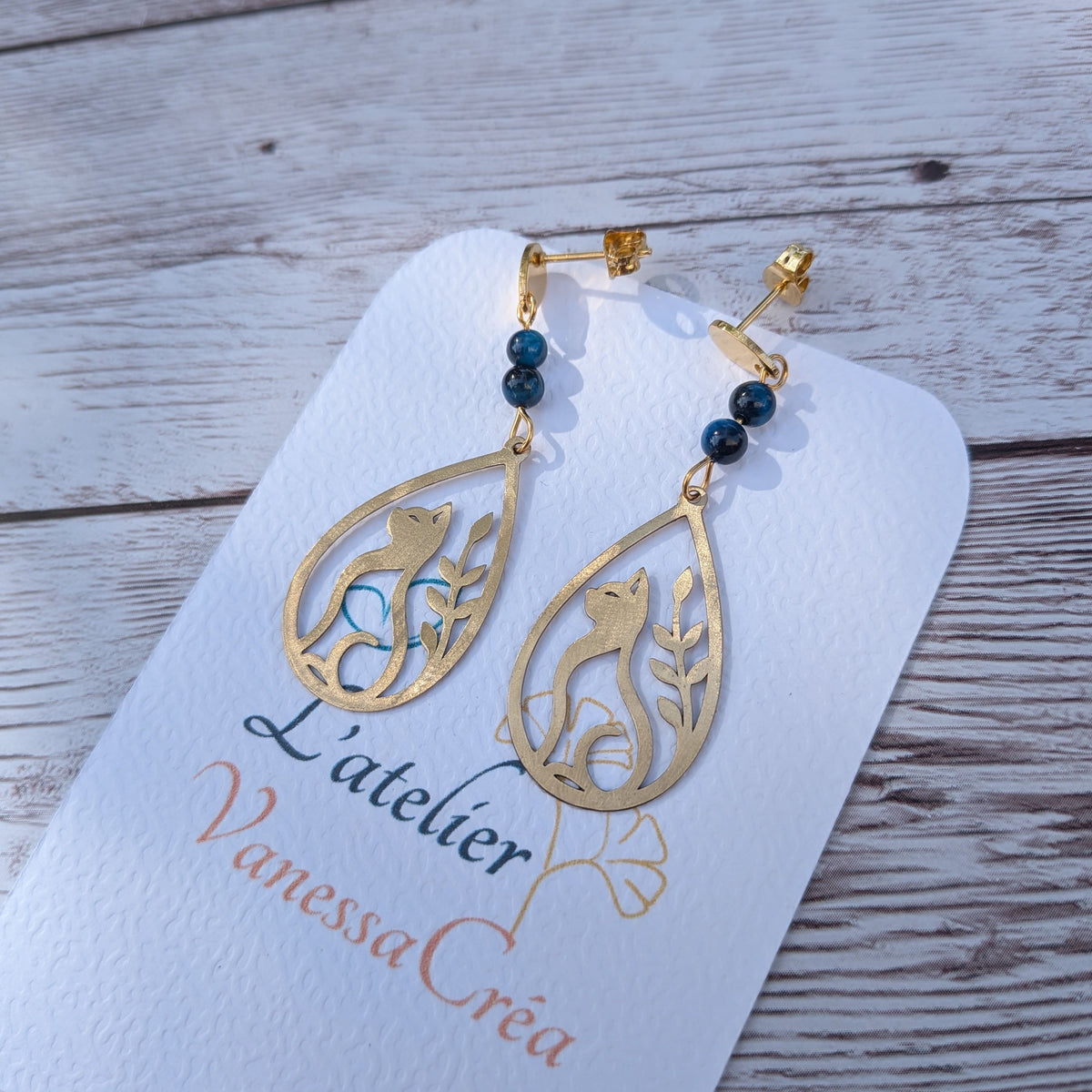 Paire de boucles d'oreilles artisanales motif chat et feuillage ajouré en laiton et œil de faucon sur support de présentation.