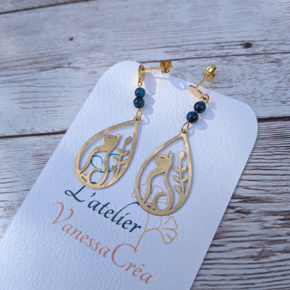 Paire de boucles d'oreilles artisanales motif chat et feuillage ajouré en laiton et œil de faucon sur support de présentation.