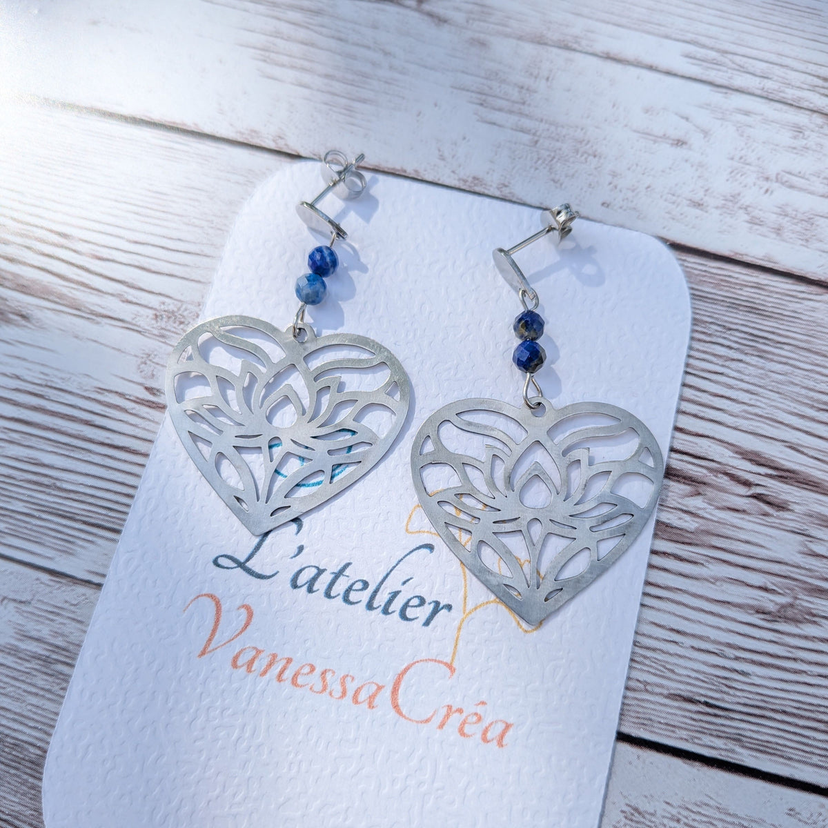 Paire de boucles d'oreilles artisanales motif cœur ajouré en acier inoxydable et lapis-lazuli sur support de présentation.