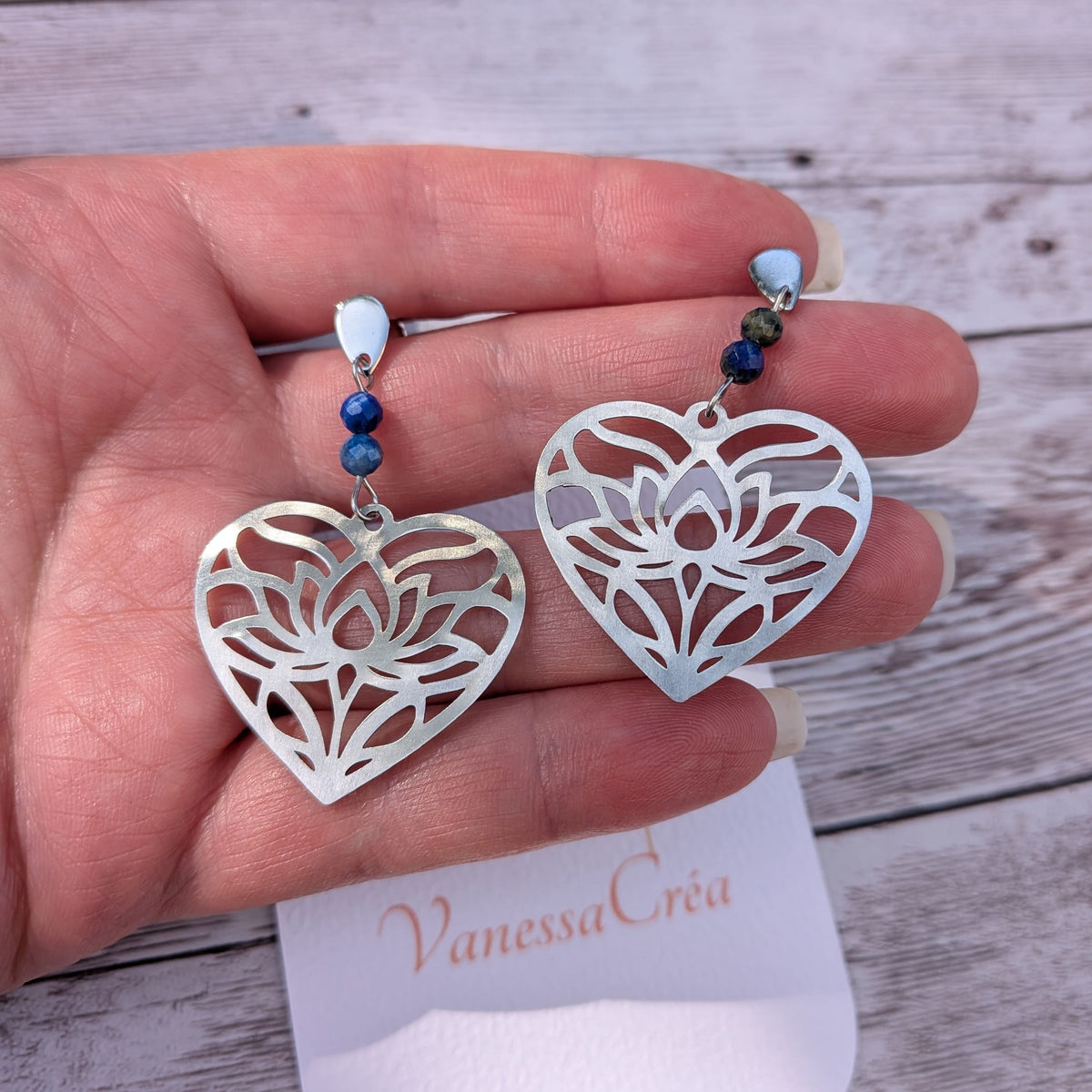 Boucles d'oreilles cœur ajouré en acier inoxydable et perles de lapis-lazuli, présentées sur la main avec la carte de l'atelier Vanessa Créa.