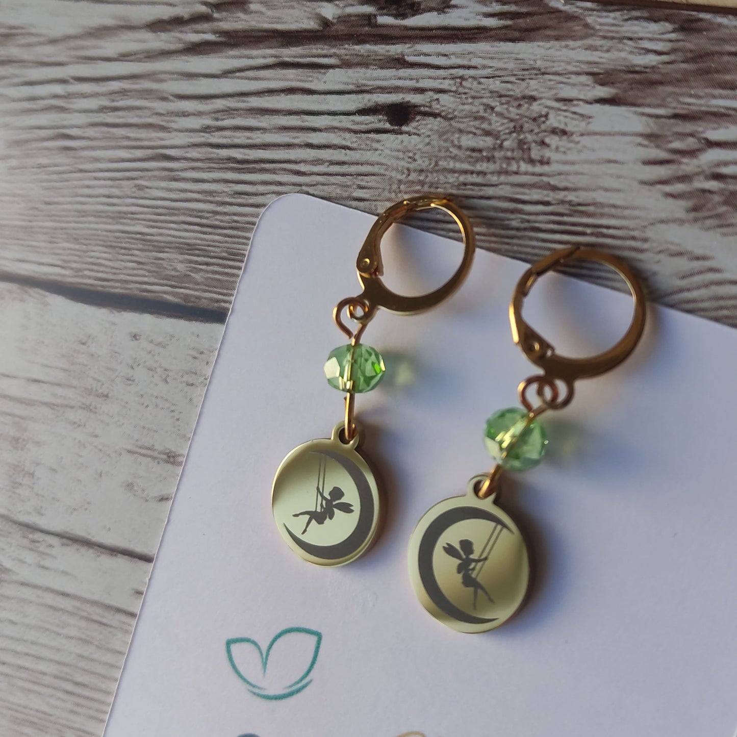Paire de boucles d'oreilles en acier inoxydable avec gravure laser d'une fée se balançant sur une lune et cristal étincelant.
