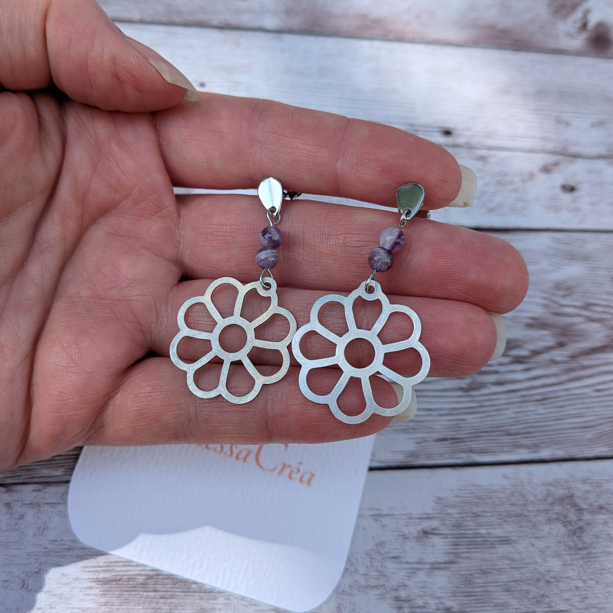 Boucles d'oreilles fleur ajourée en acier inoxydable avec perles d'améthyste de 4 mm, présentées sur la main avec la carte de l'atelier Vanessa Créa.
