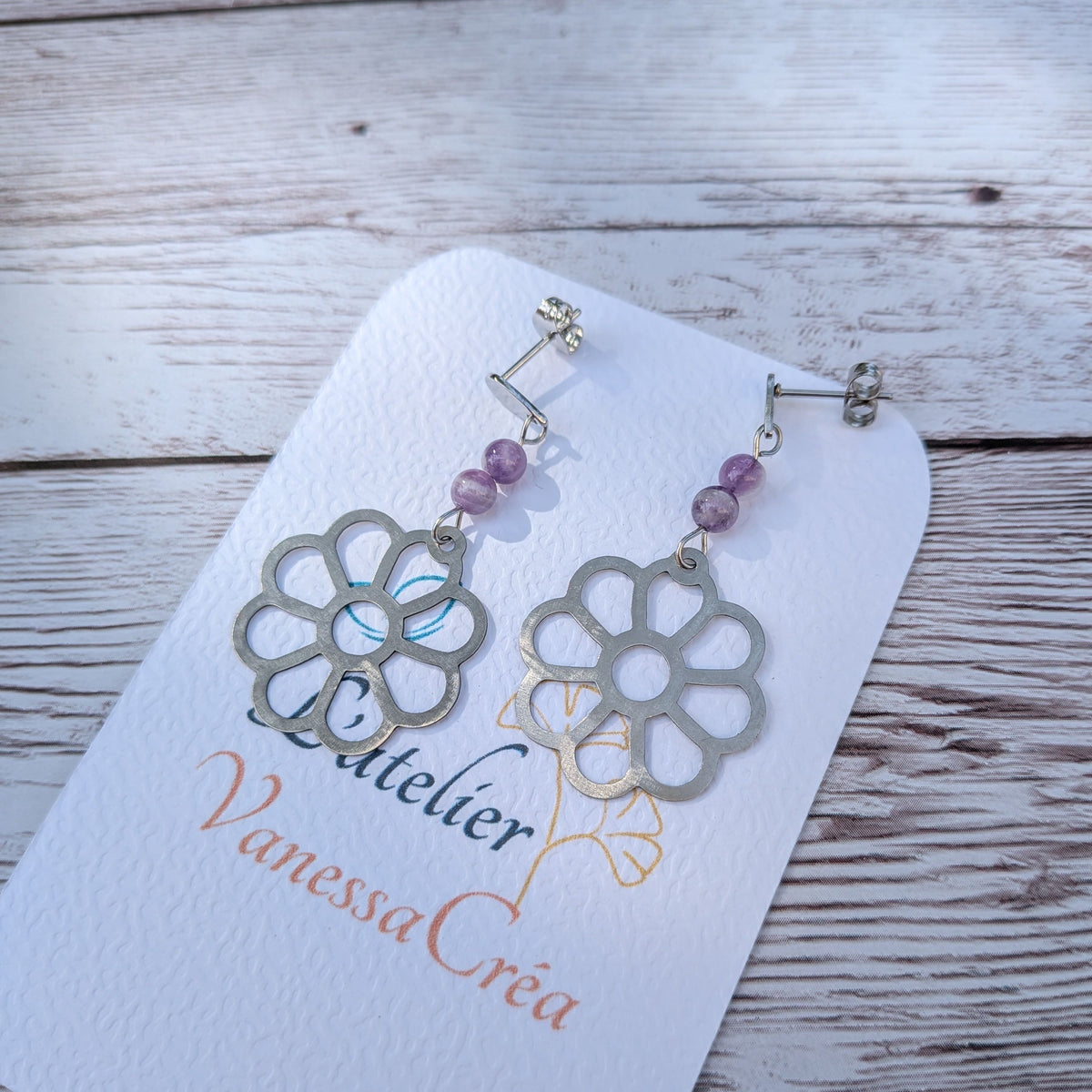 Paire de boucles d'oreilles artisanales motif fleur ajourée en acier inoxydable et améthyste sur support de présentation.
