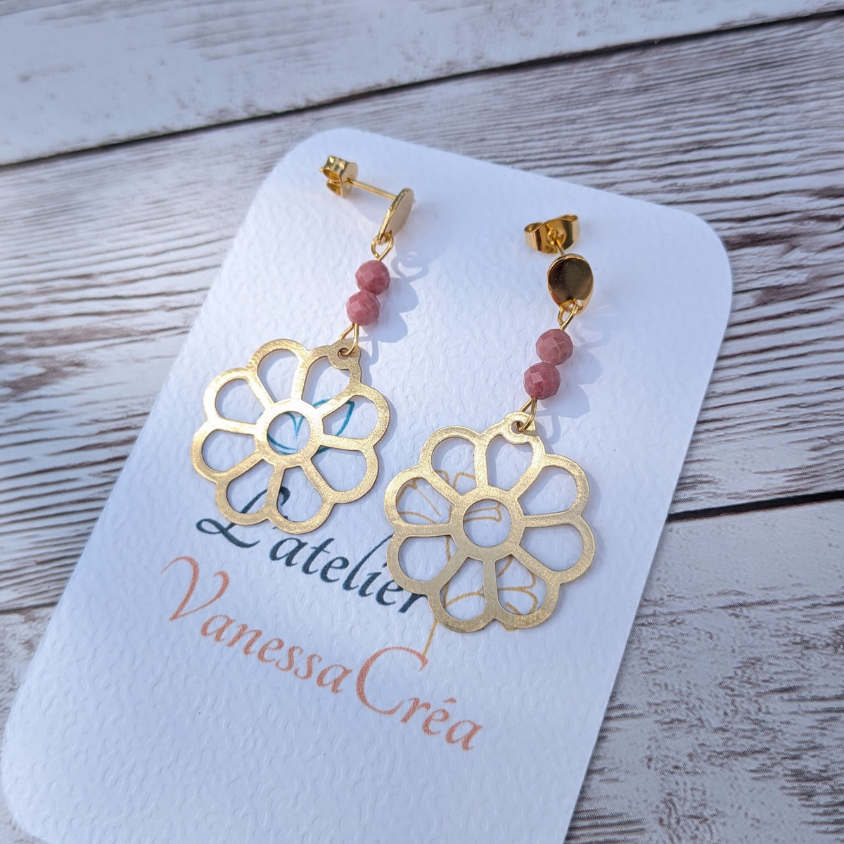 Paire de boucles d'oreilles artisanales motif fleur ajourée en laiton et rhodonite sur support de présentation.