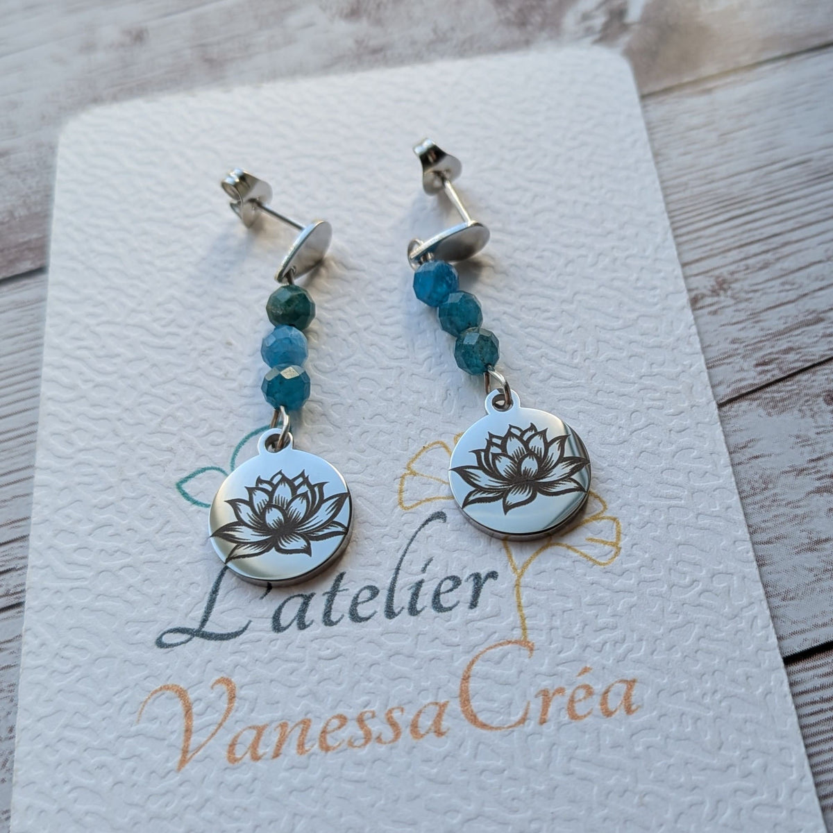 Boucles d'oreilles pendantes argentées avec fleur de lotus et apatite bleue, sur leur carte de présentation "L'atelier Vanessa Créa"
