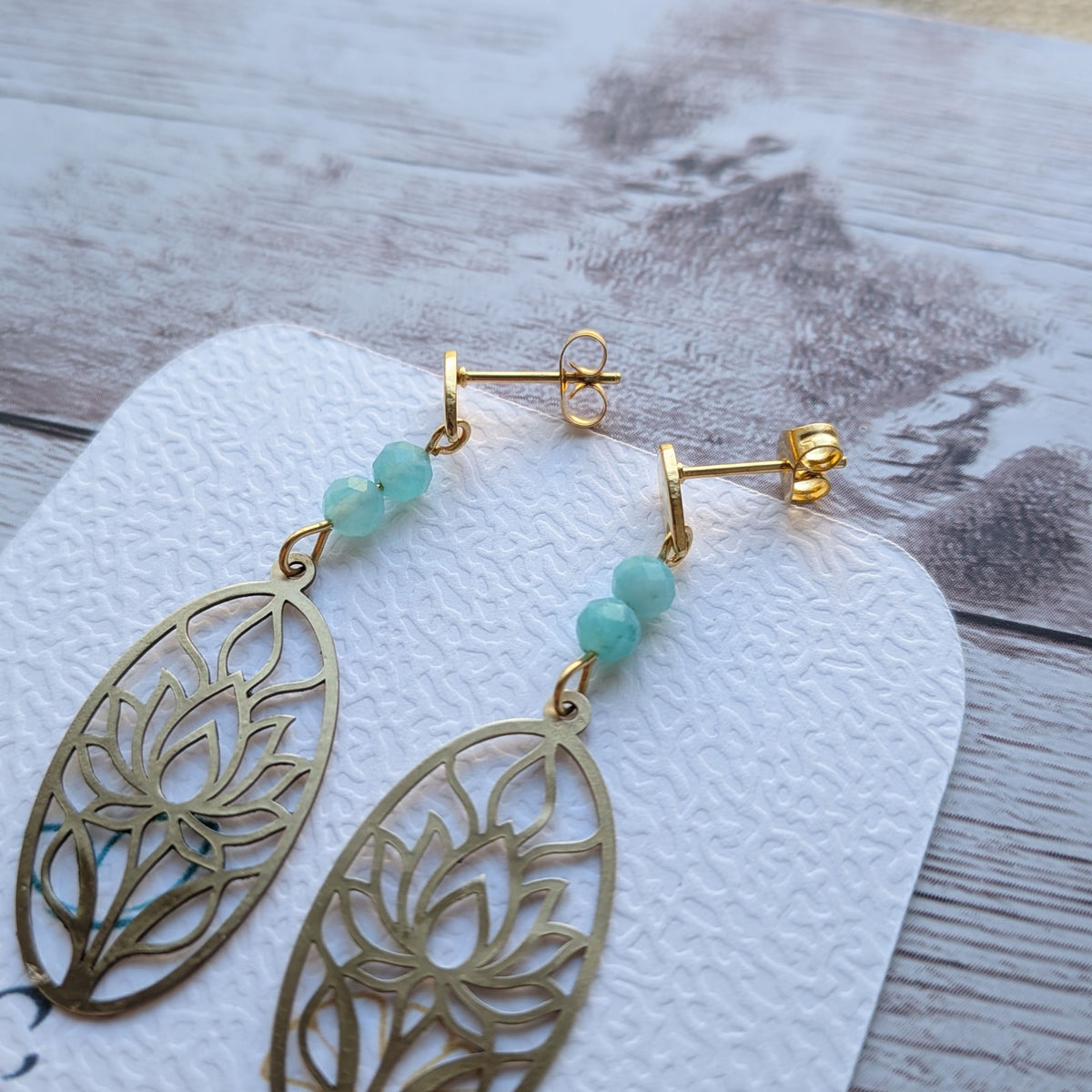 Paire de boucles d'oreilles artisanales motif lotus en laiton doré et perles d'amazonite naturelle posées sur support.