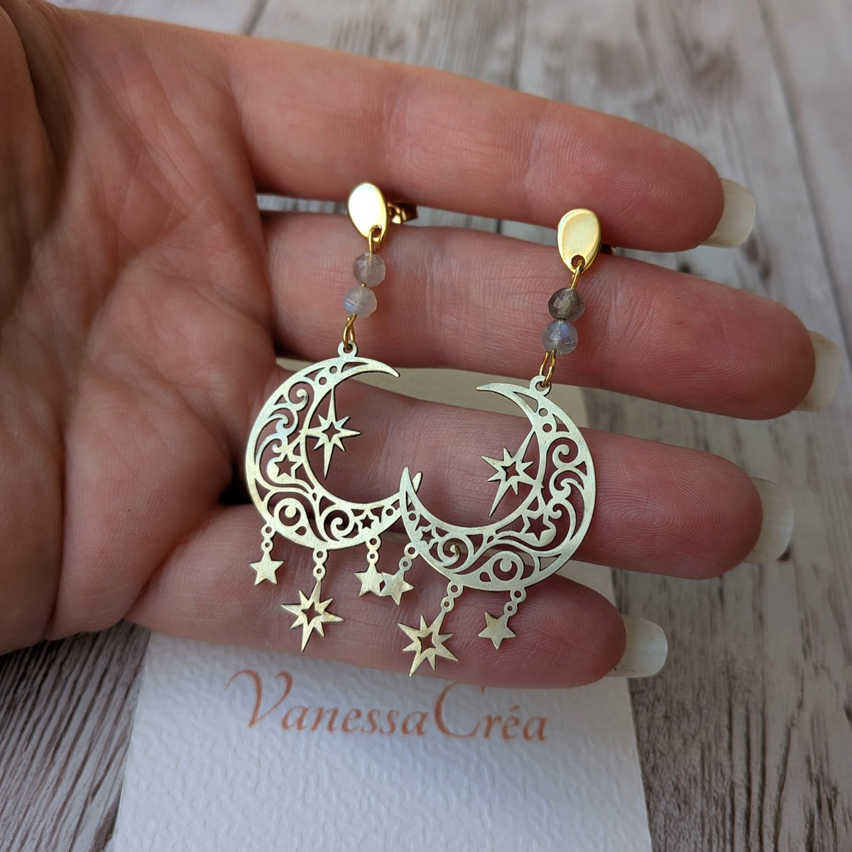 Boucles d'oreilles croissant de lune et étoiles ajourées en laiton avec perles de labradorite, présentées sur la main avec la carte de l'atelier Vanessa Créa.