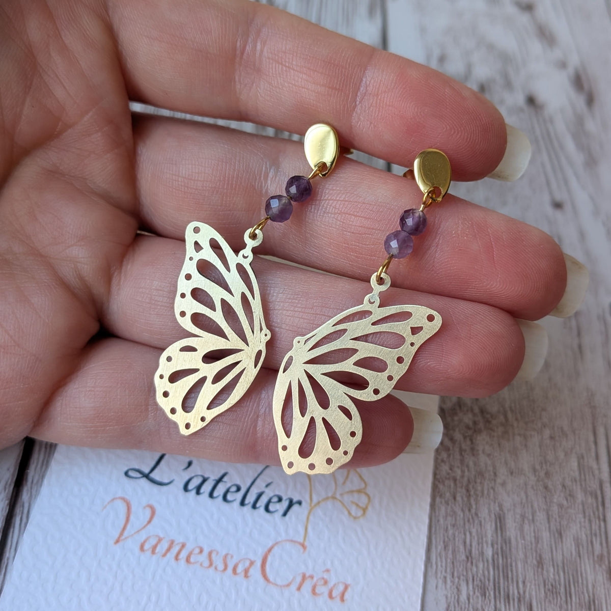 Boucles d'oreilles papillon ajouré en laiton et améthyste présentées sur fond de carte et main de l'atelier Vanessa Créa