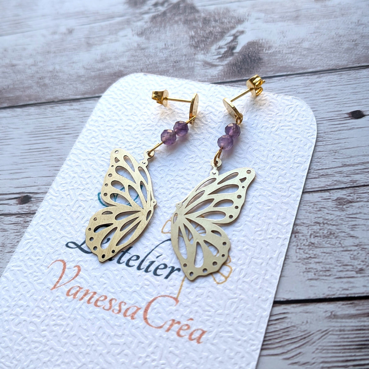 Paire de boucles d'oreilles asymétriques en laiton doré et perles d'améthyste naturelle posées sur support artisanal.