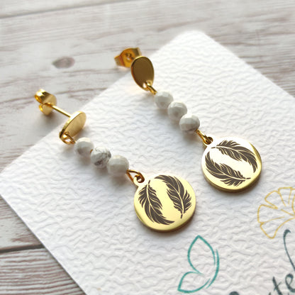 Paire de boucles d'oreilles pendantes dorées avec motif plume gravé et perles de Howlite sur carte de présentation L'atelier Vanessa Créa.