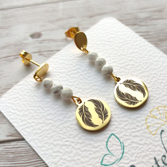 Paire de boucles d'oreilles pendantes dorées avec motif plume gravé et perles de Howlite sur carte de présentation L'atelier Vanessa Créa.