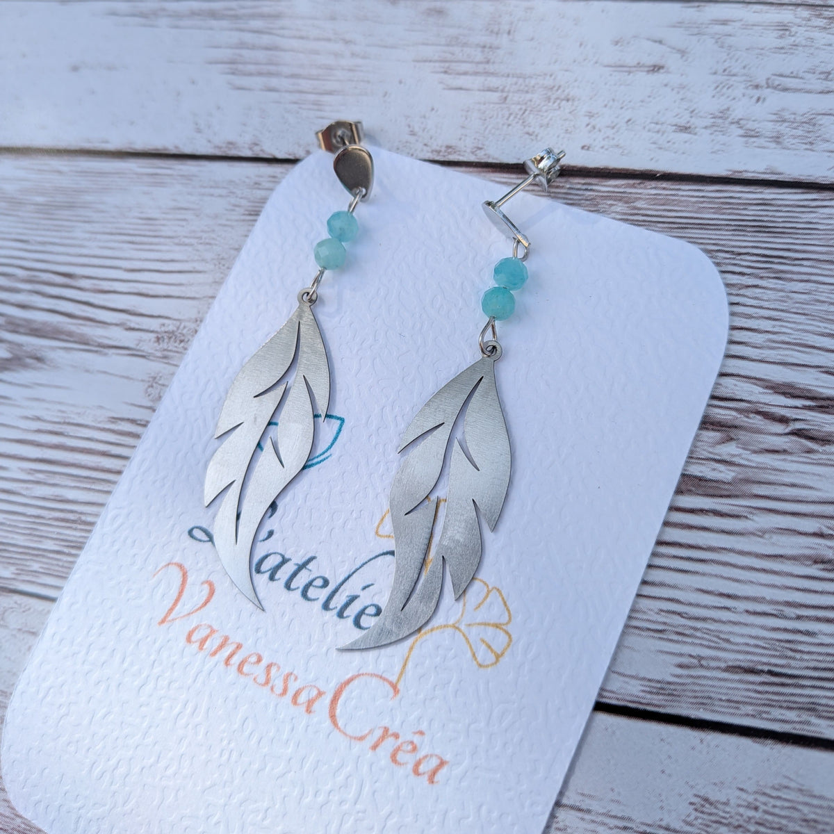 Paire de boucles d'oreilles artisanales motif plume ajourée en acier inoxydable et amazonite sur support de présentation.