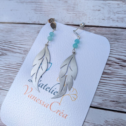 Paire de boucles d'oreilles artisanales motif plume ajourée en acier inoxydable et amazonite sur support de présentation.