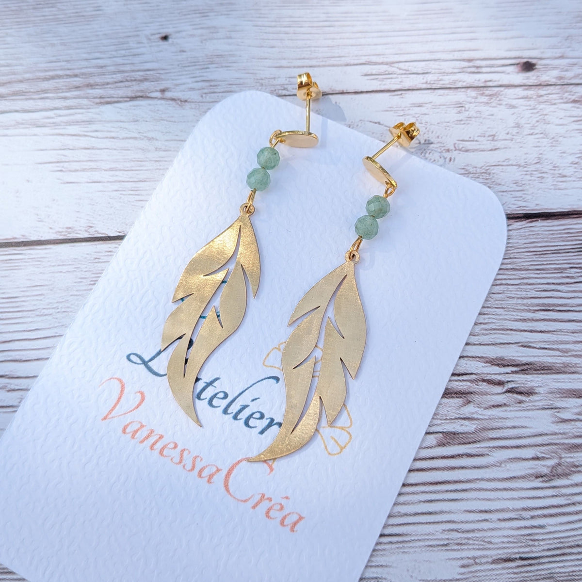 Paire de boucles d'oreilles artisanales motif plume ajourée en laiton et aventurine sur support de présentation.