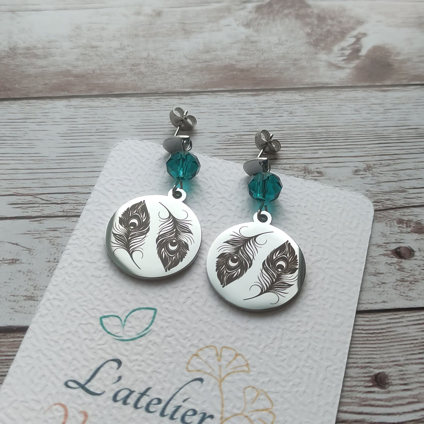 Boucles d'oreilles pendantes en acier inoxydable poli miroir avec gravure laser de deux plumes de paon et cristal à facettes.