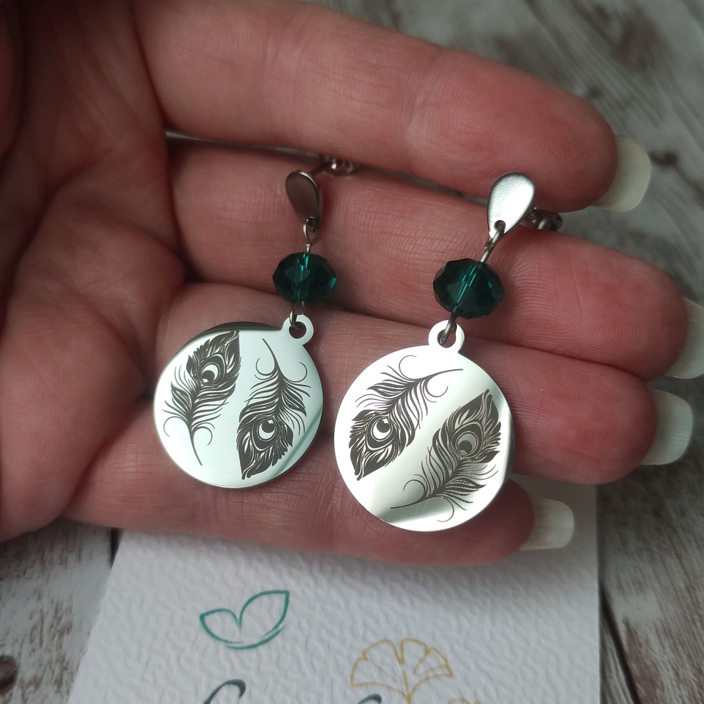 Boucles d'oreilles pendantes en acier inoxydable poli miroir avec gravure laser de deux plumes de paon et cristal à facettes.