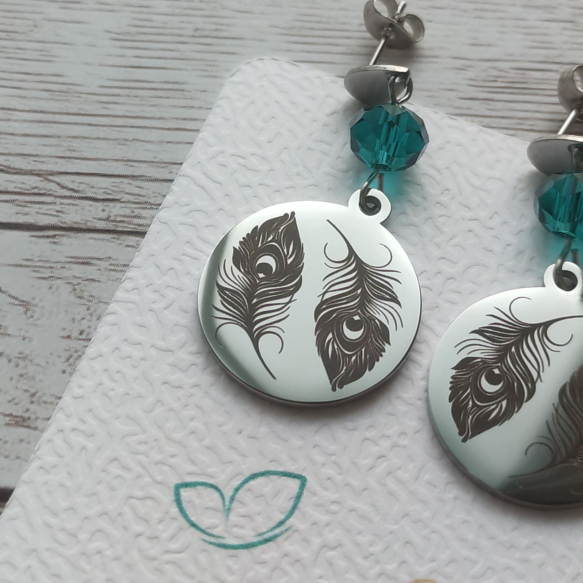 Boucles d'oreilles pendantes en acier inoxydable poli miroir avec gravure laser de deux plumes de paon et cristal à facettes.
