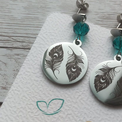 Boucles d'oreilles pendantes en acier inoxydable poli miroir avec gravure laser de deux plumes de paon et cristal à facettes.